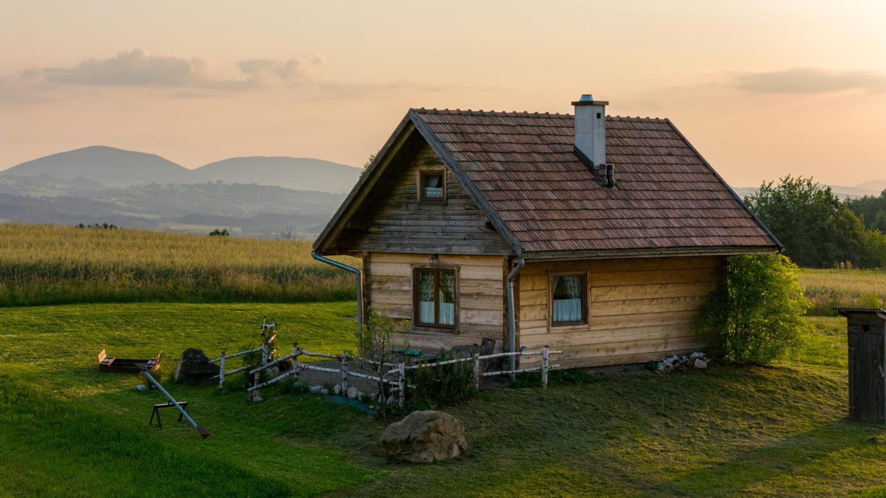 Drewniana chata na łące o zachodzie słońca, w tle pagórki i pole